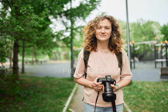 Фокусована жінка-фотограф з сучасною камерою, що стоїть в зеленому парку і дивиться на камеру — стокове фото