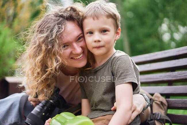 Encantada mulher amorosa abraçando menino bonito enquanto sentado no banco no parque e olhando para a câmera — Fotografia de Stock