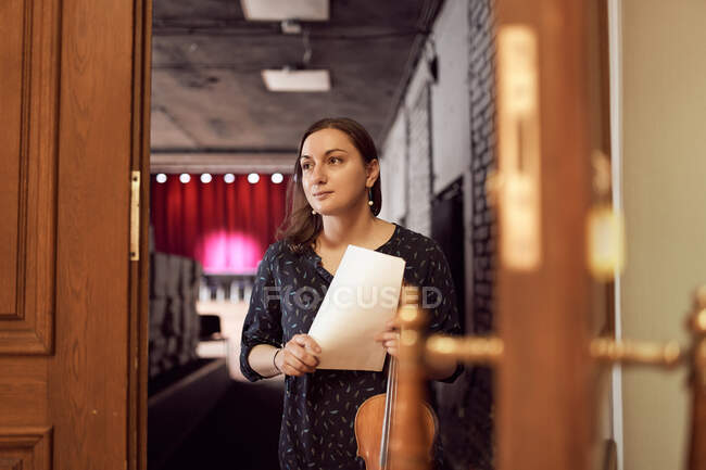 Щаслива жінка-музикантка зі скрипкою і ноткою, що стоїть біля дверей сучасного концертного залу і дивиться вбік — стокове фото