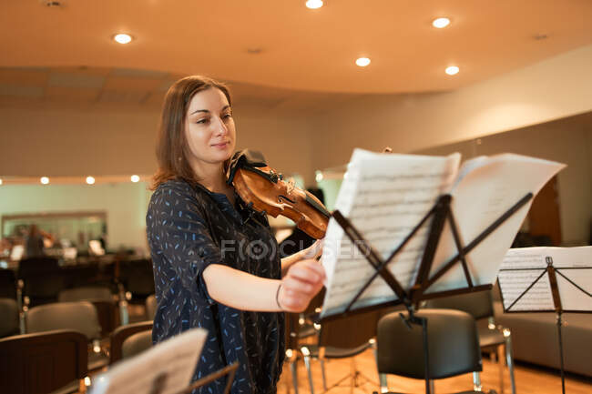 Довольная профессиональная женщина-музыкант со скрипкой, перелистывающей ноты во время репетиции в современном концертном зале — стоковое фото
