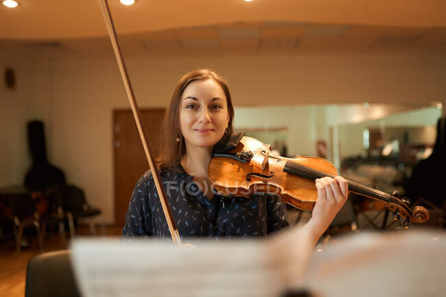 Professionelle Musikerin spielt akustische Geige und schaut während der Probe im Studio in die Kamera mit Notenblatt — Stockfoto