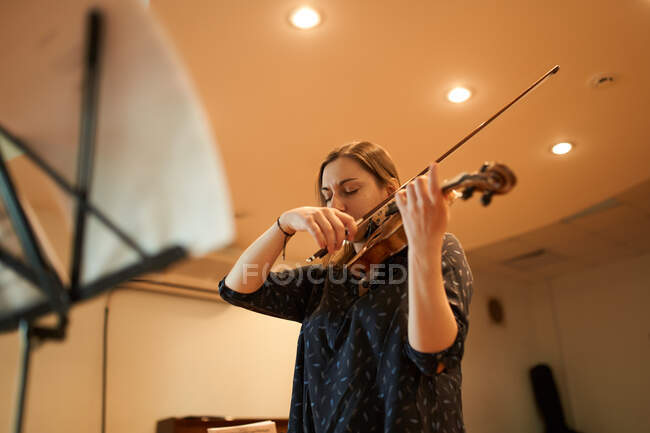 Músico profesional enfocado tocando violín acústico con los ojos cerrados con hoja de música durante el ensayo en estudio - foto de stock