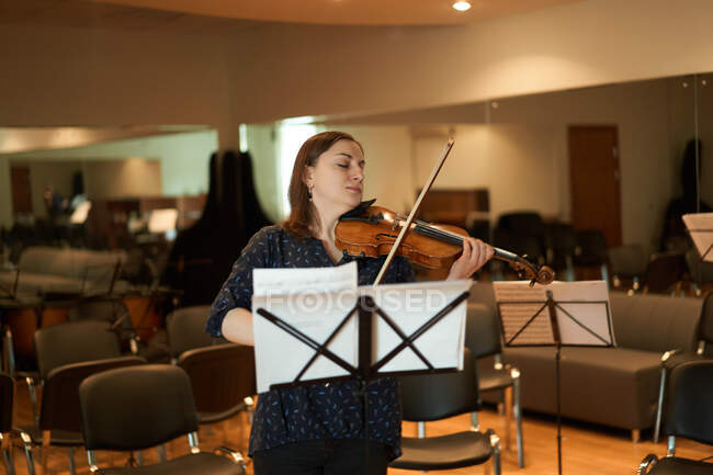 Focused professional female musician playing acoustic violin with eyes closed with music sheet during rehearsal in studio — Stock Photo