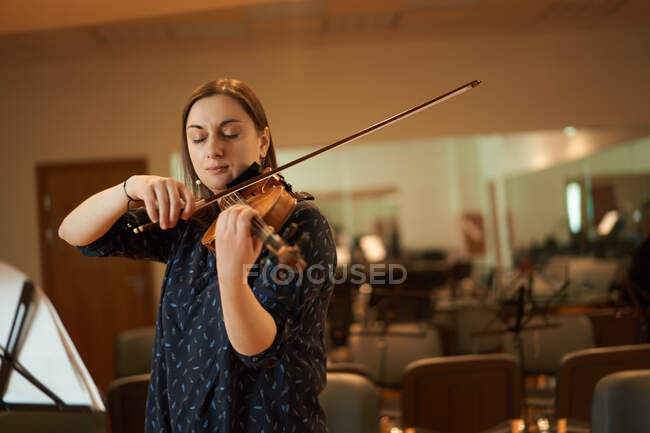 Focused professional female musician playing acoustic violin with eyes closed with music sheet during rehearsal in studio — Stock Photo