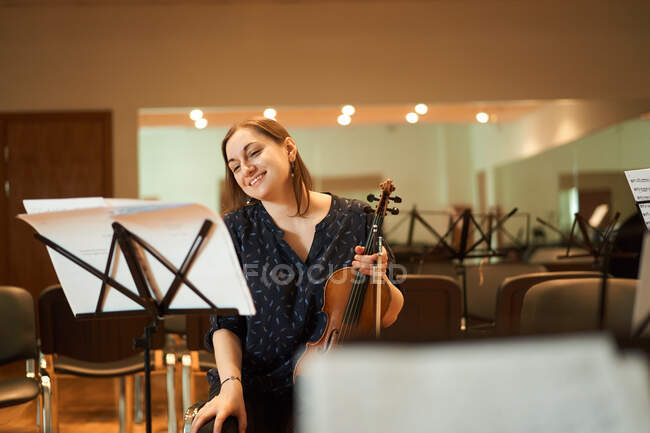 Músico feminino profissional alegre tocando violino acústico e olhando para a folha de música durante o ensaio em estúdio — Fotografia de Stock