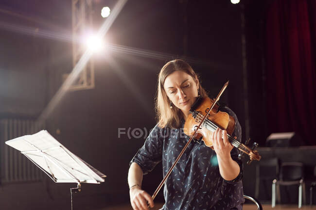 Focused female musician playing violin near stand with sheet music in bright lights in concert hall — Stock Photo