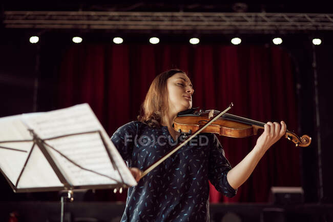 Focused female musician playing violin with eyes closed near stand with sheet music in bright lights in concert hall — Stock Photo
