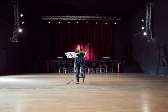 Músico femenino enfocado tocando el violín cerca del stand con partituras en luces brillantes en la sala de conciertos - foto de stock