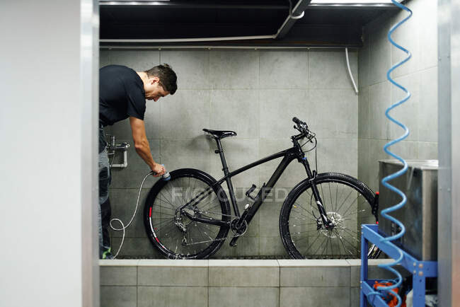 Vista lateral de la caja de engranajes de limpieza mecánica masculina de rueda de bicicleta con agua en el taller - foto de stock