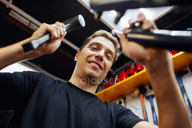 From below of delighted male mechanic with hammer fixing bike in modern garage — Stock Photo
