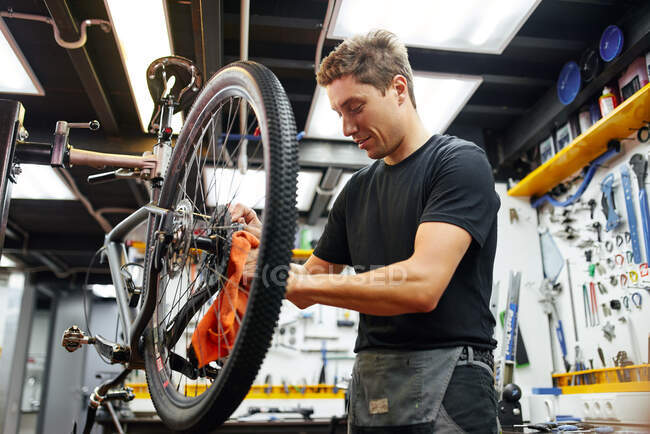 Marco masculino enfocado de la bici del limpiamiento principal con el trapo mientras que trabaja en taller moderno de la reparación - foto de stock