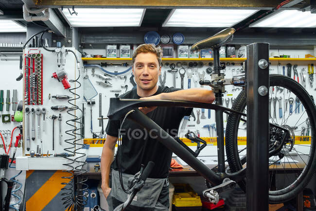 Happy male mechanic smiling and looking at camera while leaning on bike under repair against wall with tools in garage — Stock Photo