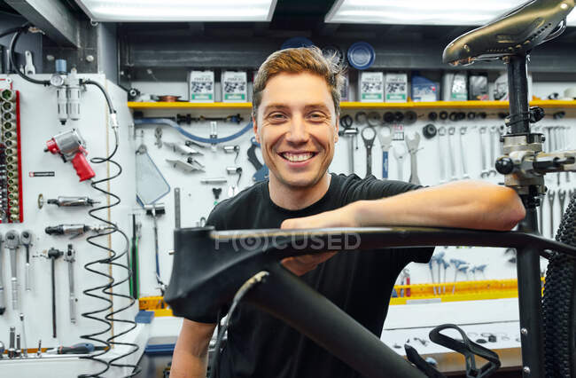 Joyeux mécanicien masculin souriant et regardant la caméra tout en s'appuyant sur le vélo en réparation contre le mur avec des outils dans le garage — Photo de stock