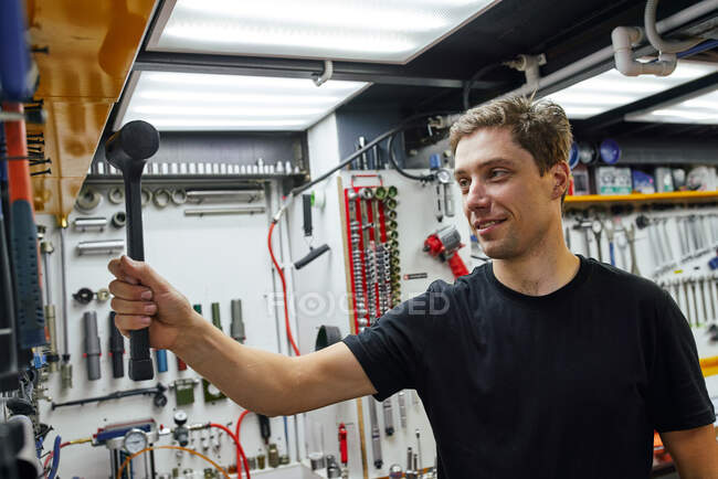 Erwachsener Mann in schwarzem T-Shirt lächelt und holt Hammer von Wand, während er in professioneller Werkstatt arbeitet — Stockfoto