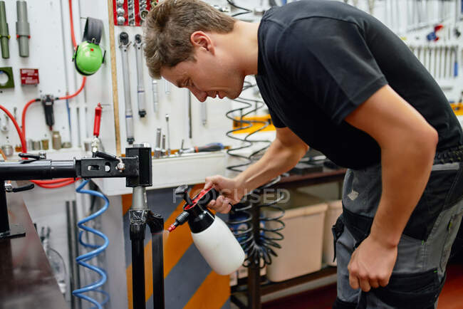 Vue latérale de l'homme adulte pulvérisation d'eau et nettoyage fourche de vélo attaché au rack dans l'atelier de réparation moderne — Photo de stock