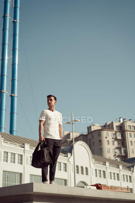 Low angle of male athlete with sports bag and in activewear standing on stairs in city and looking away — Stock Photo