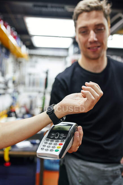 М'який фокус чоловічої механіки з терміналом отримання безконтактних платежів від клієнта врожаю з розумним годинником під час роботи в гаражі — стокове фото