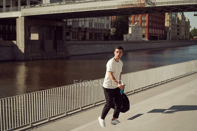 Male athlete with sports bag walking upstairs on embankment in city in summer — Stock Photo