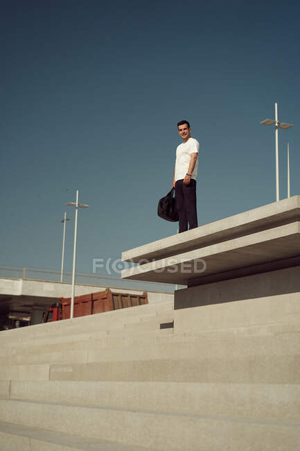 Low angle of delighted male athlete with sports bag and in activewear standing on stairs in city and looking at camera — Stock Photo