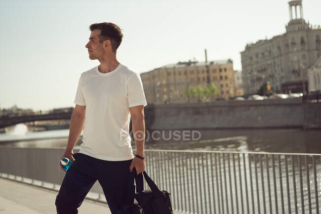 Zufriedener männlicher Athlet mit Sporttasche und Wasserflasche steht im Sommer auf einem Damm in der Stadt — Stockfoto