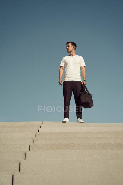 Низкий угол у спортсмена с спортивной сумкой и в спортивной одежде, стоящего на лестнице в городе и смотрящего в сторону — стоковое фото