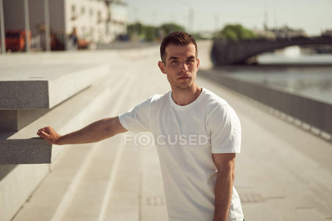 Handsome male in white t shirt standing on promenade in city on sunny day and looking at camera — Stock Photo