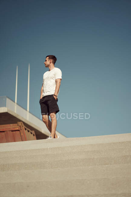 Angolo basso di atleta in forma maschile in abbigliamento sportivo in piedi sulle scale in città nella giornata di sole e guardando altrove — Foto stock
