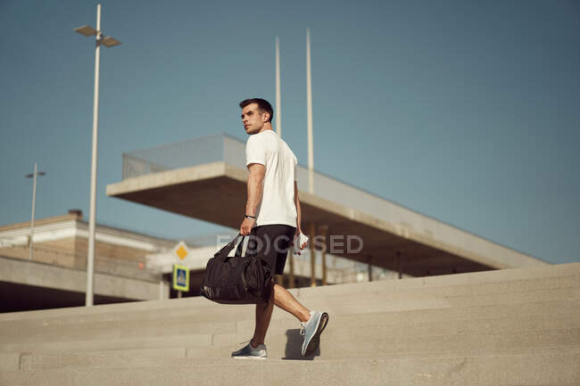 Low angle of male athlete with sports bag and in activewear walking on stairs in city and looking away — Stock Photo
