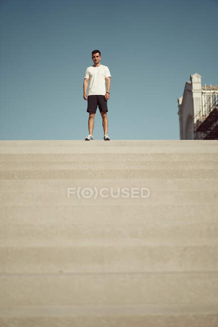 Низький кут підгонки спортсмена-чоловіка в спортивному одязі, що стоїть на сходах у місті в сонячний день і дивиться на камеру — стокове фото