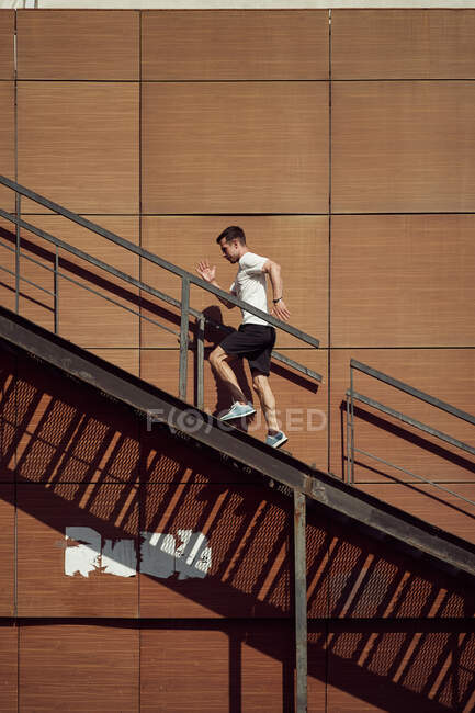 Side view of energetic male athlete running up metal staircase near building during training in summer — Stock Photo