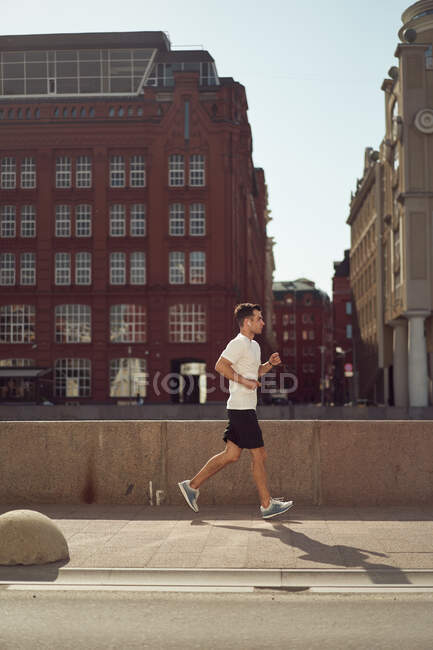 Vista lateral de un fuerte atleta masculino corriendo por la calle durante el entrenamiento activo en la ciudad en verano - foto de stock