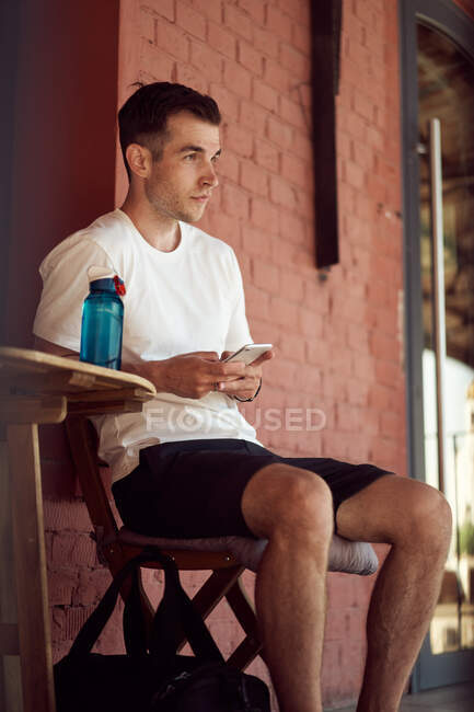 Male athlete sitting in street cafe with bottle of water and using mobile phone after workout in city — Stock Photo