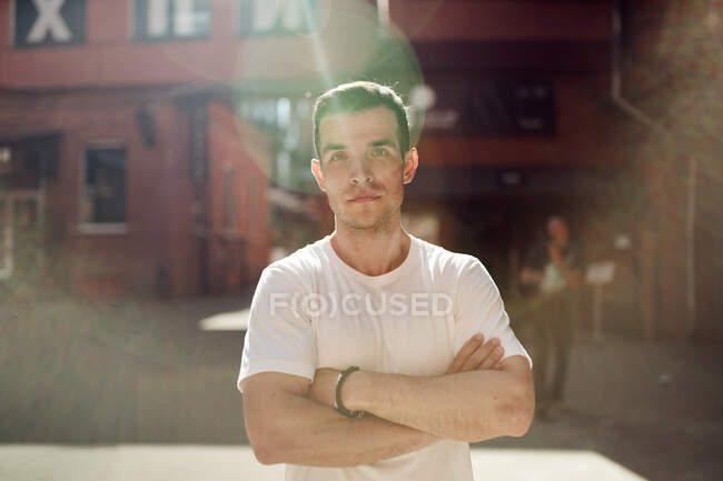 Determinado bonito masculino de pé com cruzado braços no cidade rua no ensolarado dia e olhando para a câmera — Fotografia de Stock