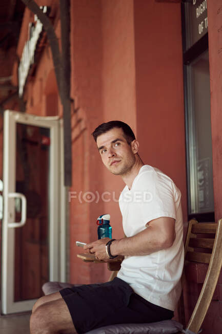 Male athlete sitting in street cafe with bottle of water and using mobile phone after workout in city — Stock Photo