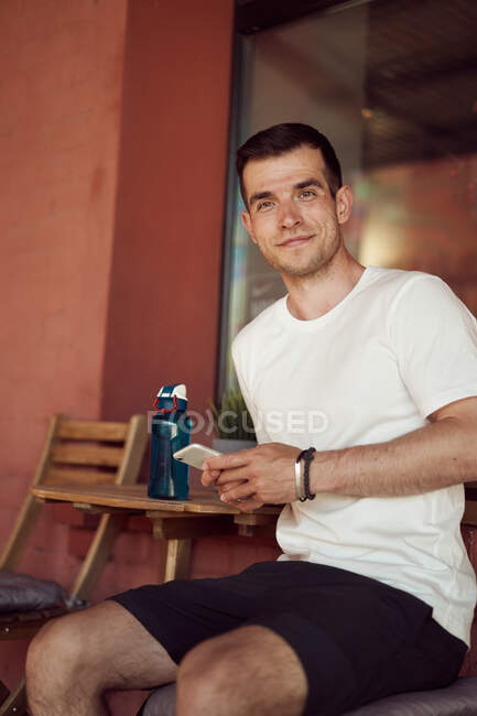 Lächelnder männlicher Athlet sitzt mit Wasserflasche im Straßencafé und benutzt Handy nach dem Training in der Stadt und schaut in die Kamera — Stockfoto