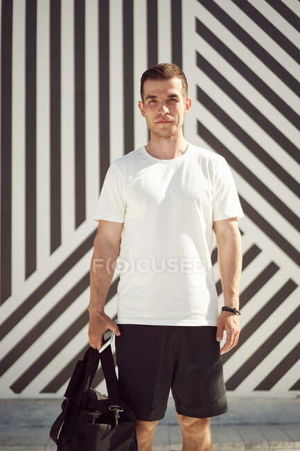 Серйозний спортсмен в активному одязі і з сумкою, що стоїть на вулиці влітку і дивиться на камеру — стокове фото