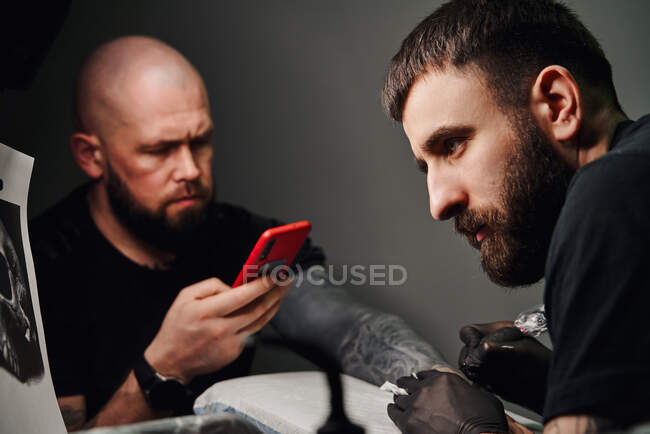 Серьезный мужчина татуировщик с помощью татуировки машины, делая татуировку на руке парня с телефоном в студии легкой татуировки — стоковое фото