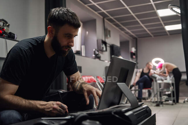 Thoughtful adult guy in casual outfit with tattoos sketching on computer in bright tattoo studio near tattooist making tattoo for client — Stock Photo