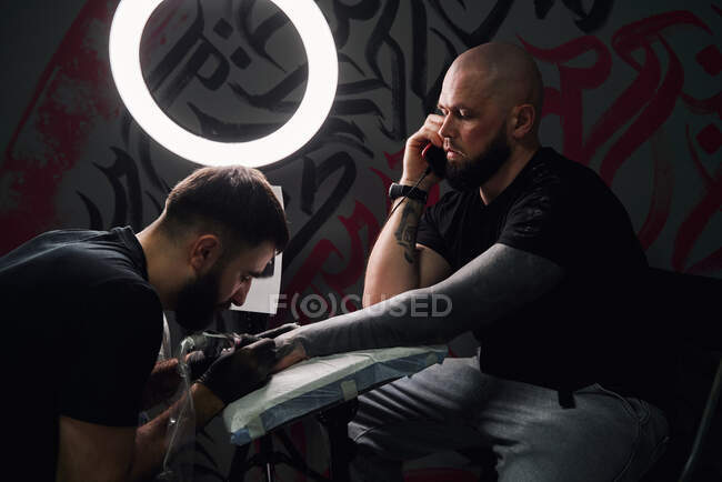 Серйозний чоловічий майстер татуювання з використанням татуювання машини під час створення татуювання на руці хлопця з телефоном у студії легких татуювань — стокове фото