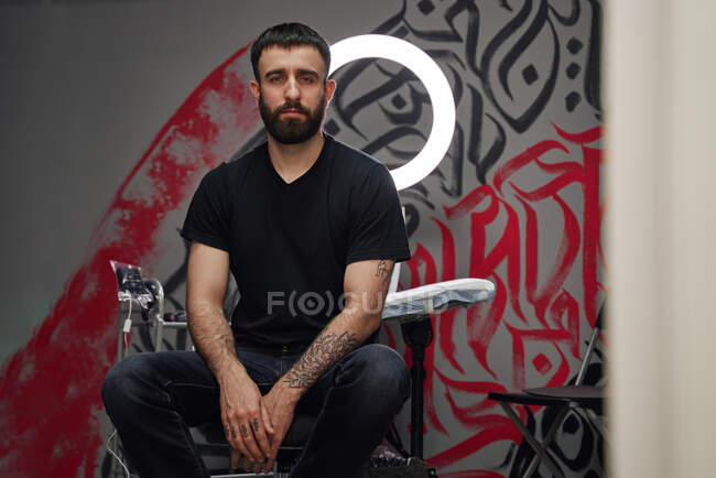 Tatuaje masculino adulto barbudo reflexivo en ropa casual sentado en el taburete cerca de la lámpara del anillo y los instrumentos mientras mira a la cámara en el salón de tatuajes de luz con graffiti en la pared - foto de stock