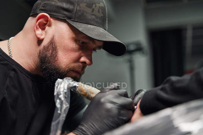 Tatuagem masculina concentrada em luvas fazendo tatuagem na mão do cliente enquanto usa máquina de tatuagem profissional no estúdio de tatuagem moderno — Fotografia de Stock