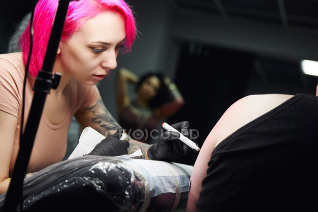 Серьезный молодой мастер татуировки в перчатках рисует татуировку на плече клиента маркером перед тем, как сделать татуировку в яркой студии — стоковое фото