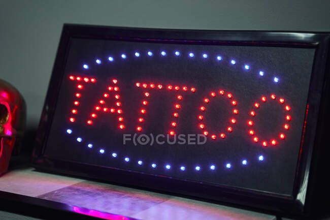 Panneau lumineux avec tatouage mot rouge autour des néons bleus sur l'étagère à côté de faux crâne doré et respirateur près du mur noir dans studio de tatouage léger — Photo de stock
