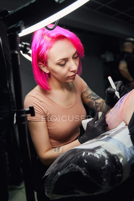 Mestre de tatuagem jovem sério em luvas desenho tatuagem no ombro do cliente com marcador antes de fazer tatuagem em estúdio brilhante — Fotografia de Stock