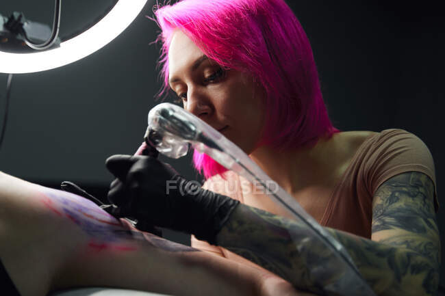 Serious tattoo master with pink hair in gloves using professional tattoo machine while making tattoo on shoulder of client in modern tattoo salon — Stock Photo