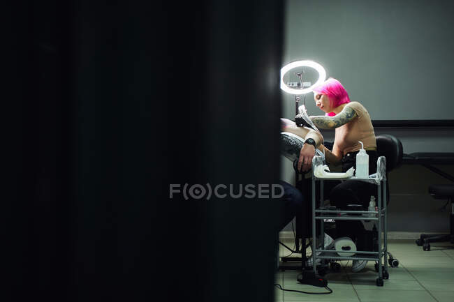 Серйозний майстер татуювання з рожевим волоссям в рукавичках, використовуючи професійну татуювальну машину, роблячи татуювання на плечі клієнта в сучасному татуювальному салоні — стокове фото