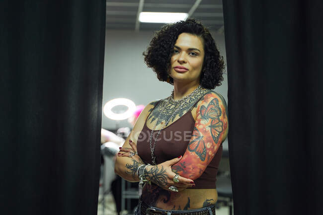 Серьезная взрослая женщина в повседневной одежде с татуировками, стоящая в салоне легких татуировок, глядя на камеру со скрещенными руками — стоковое фото