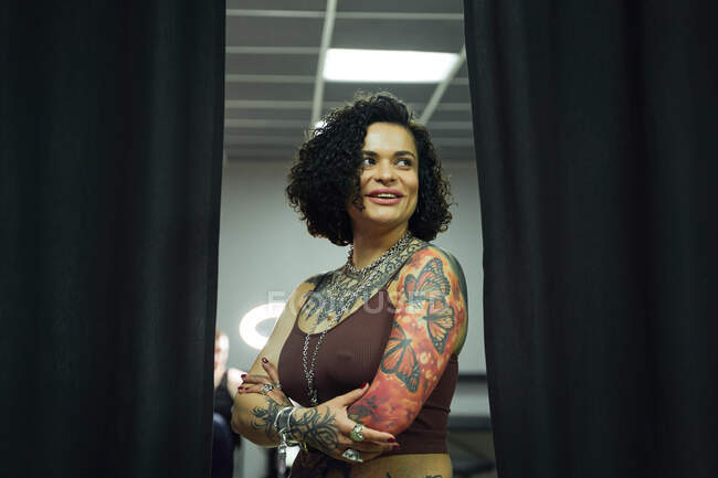 Позитивна доросла жінка в повсякденному одязі з татуюваннями, що стоять у легкому татуюванні салону, дивлячись в сторону схрещеними руками — стокове фото