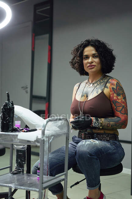 Серьезная взрослая женщина в повседневной одежде с татуировками, сидящая в салоне легких татуировок, глядя в камеру — стоковое фото