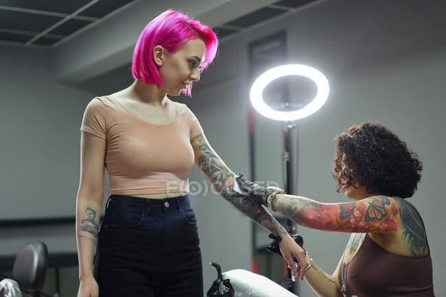 Side view tattooist in latex gloves wiping freshly made tattoo on arm of female client with napkin while working in salon — Stock Photo